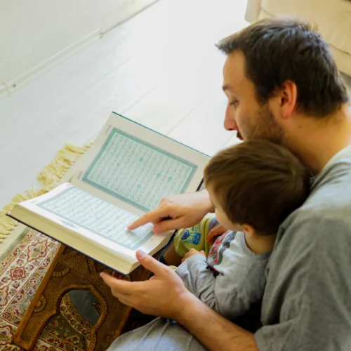 A father and his young son sitting together on the floor studying the Quran.